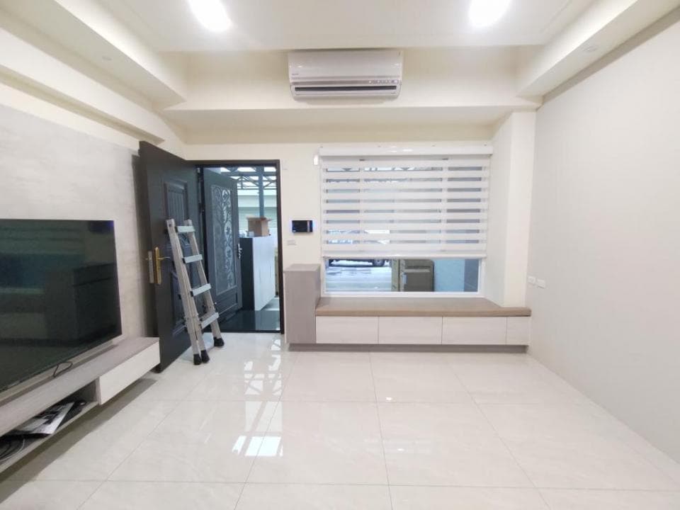 1) 照片顯示一個現代化住宅的客廳,地板鋪設白色磁磚,牆壁為白色,客廳中央放置一台電視,旁邊有一個木質電視櫃,客廳的右邊有一扇落地窗,窗戶裝有白色百葉窗,窗戶下方有一個木質窗台,窗台下方有一個木質櫃子,客廳的左邊有一扇門,門旁邊有一台梯子