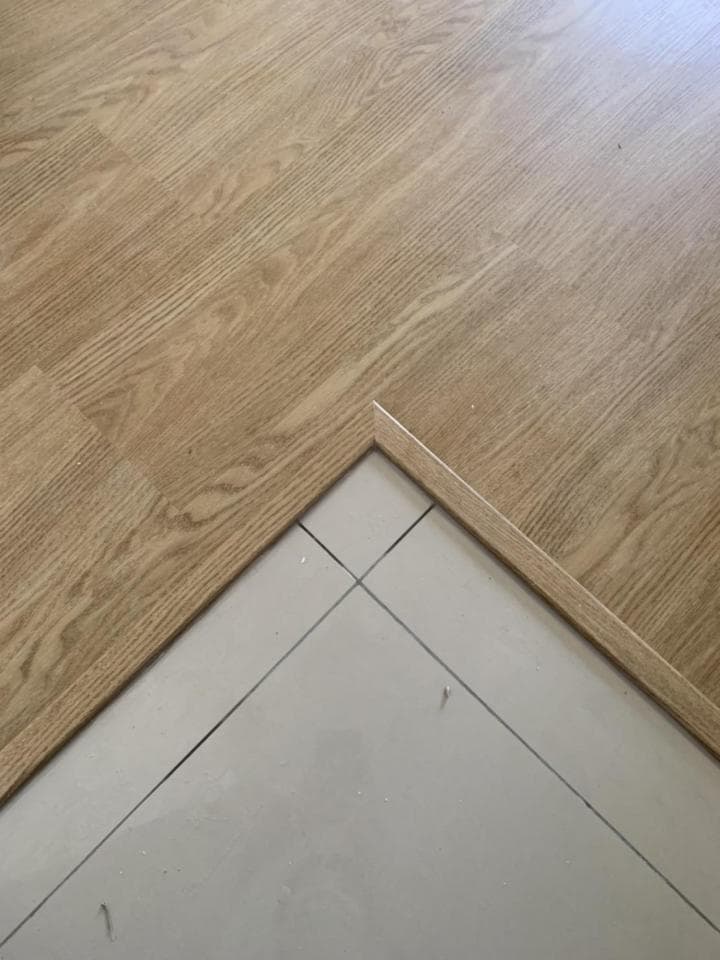 這張照片是兩塊木地板的接縫處,兩塊木地板都是淺褐色,木紋清晰可見。地板的接縫處用白色膠帶固定,膠帶上還有一些黑色的線條。
