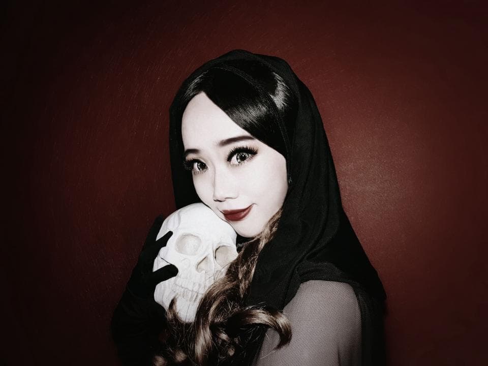 這張照片是一個穿著黑色斗篷的年輕女人,她手裡拿著一個白色的頭骨,並對著鏡頭微笑。她的妝容很精緻,眼神充滿了神秘感。