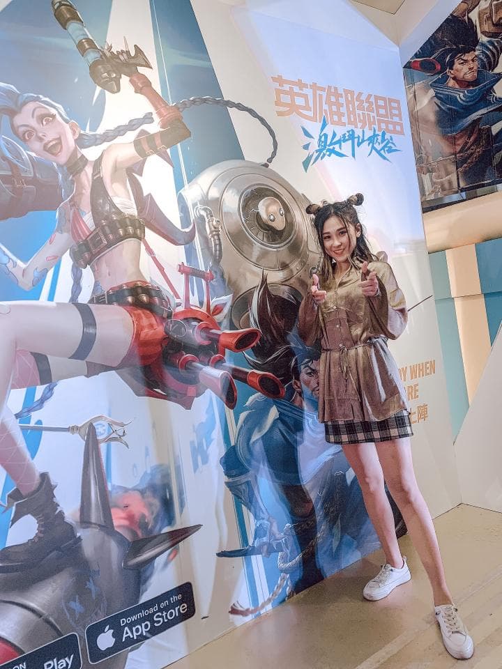 這張「英雄聯盟」的照片是一個年輕女性站在牆壁前,牆壁上畫著遊戲的標誌和角色。她穿著短裙和短袖上衣,手裡拿著手機,看起來很興奮。