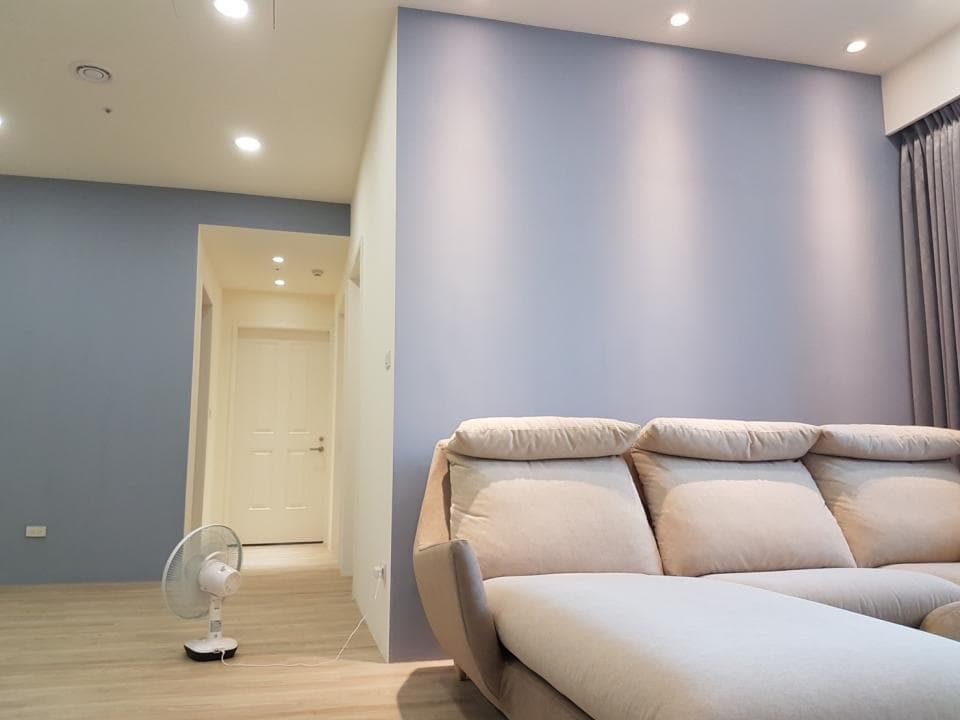 1) 照片顯示一個客廳,裡面有一張白色沙發,靠牆擺放,沙發旁有一個白色電風扇,房間牆壁為淺藍色,牆壁上裝有兩個白色燈泡