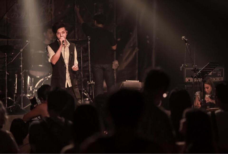 這張「演唱會」的照片是一個男歌手在舞臺上唱歌,他穿著黑色的西裝外套,拿著麥克風唱歌,他後面有一個樂隊在伴奏。舞臺上還有很多觀眾在觀看演出。