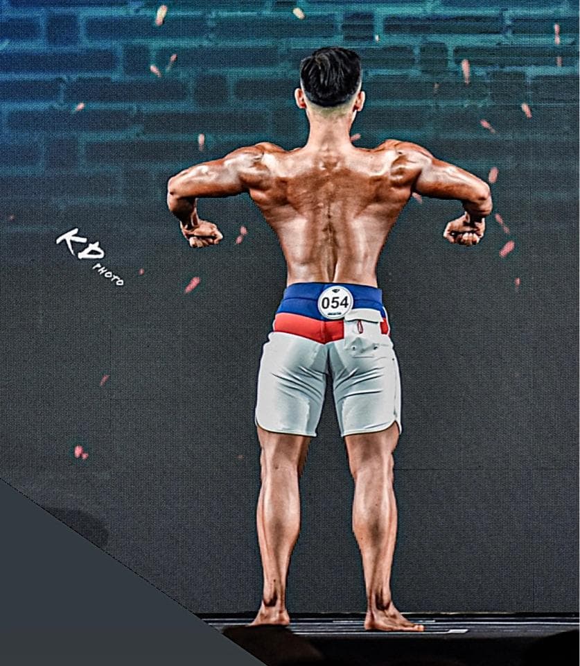 這張照片是一個健美比賽的後臺,照片中有一位健美運動員展示他精壯的背部肌肉。他穿著白色短褲,腰間繫著藍紅色的比賽腰帶,腰帶上印著號碼 054。