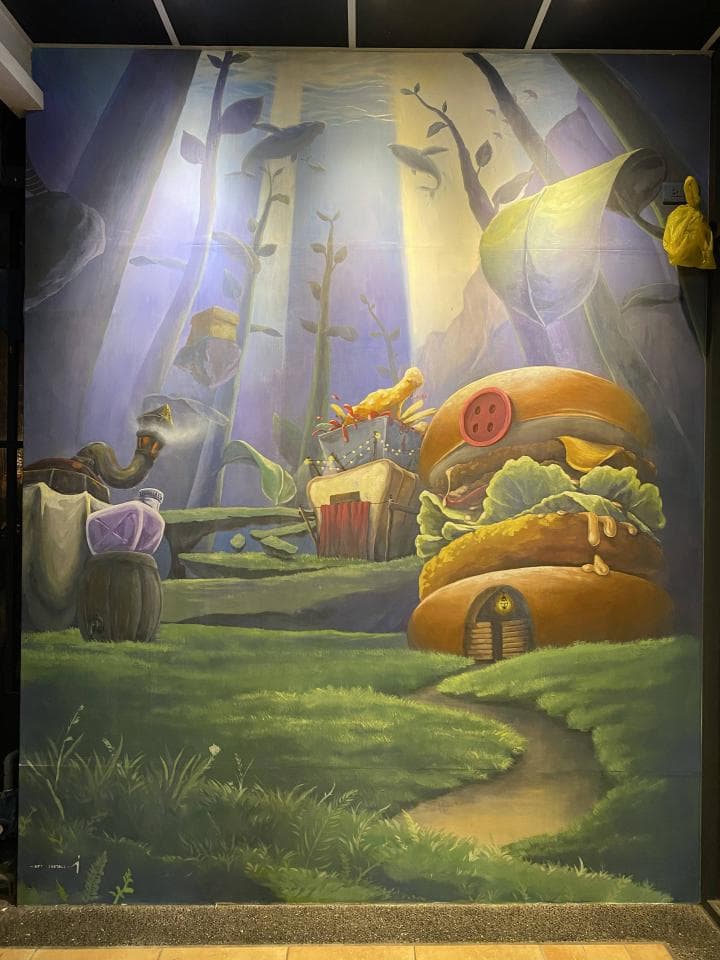 這張「牆壁上繪畫」的照片是一個童話般的森林場景,畫面中有一個巨大的漢堡,還有森林、樹木和一個可愛的卡通人物。