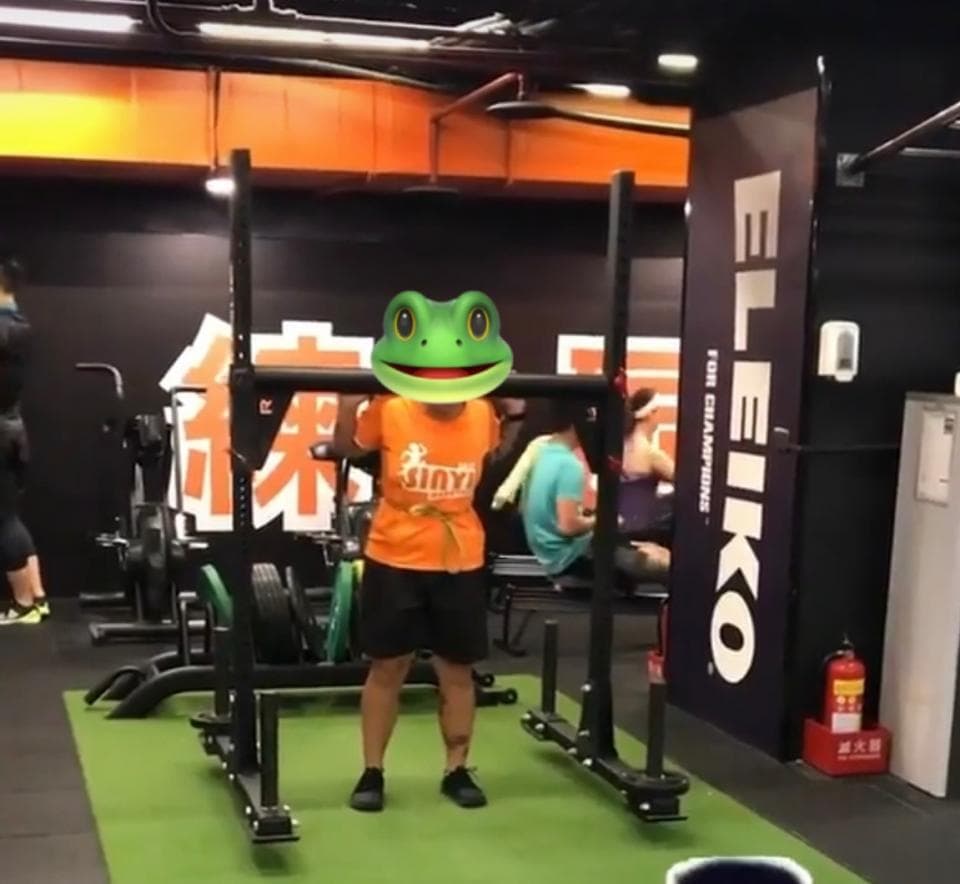 這張照片是一個健身房,照片中最重要的部分是穿著綠色青蛙頭的男子。他正在使用一個黑色的健身器械,這個器械看起來很沉重,他正在努力地舉起它。男子表情堅定,似乎很努力。