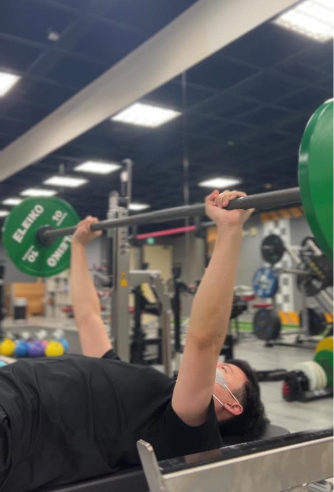 這張照片是一個男人在健身房裡做臥推。他戴著口罩,正在舉起一個重量很大的槓鈴。