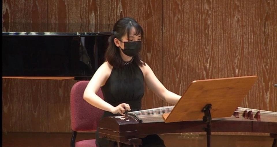 這張照片是一位年輕女子演奏古箏。她戴著黑色口罩,專注地看著古箏。她的手指靈活地在琴絃上跳動,演奏著優美的旋律。