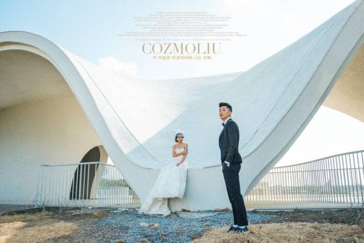 這張婚紗照是一對新人站在白色建築前,新娘穿著白色婚紗,新郎穿著黑色西裝。