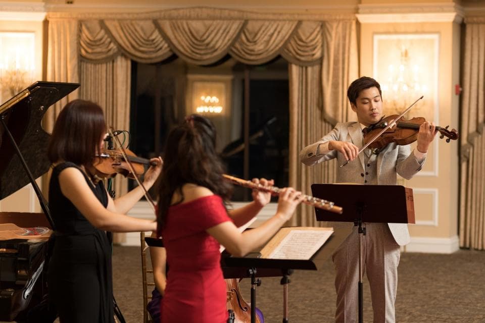 這張「四個音樂家在演奏」的照片是:一位男士拉著小提琴,一位女士吹著長笛,另一位女士拉著小提琴,還有鋼琴放在她們旁邊。