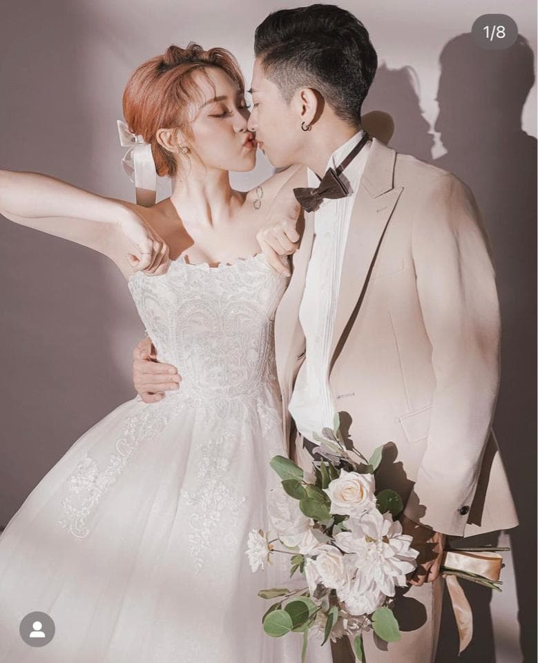 這張婚紗照是一對新人正在親吻,新娘穿著白色婚紗,新郎穿著米色西裝,他們手裡拿著一束白色花束。