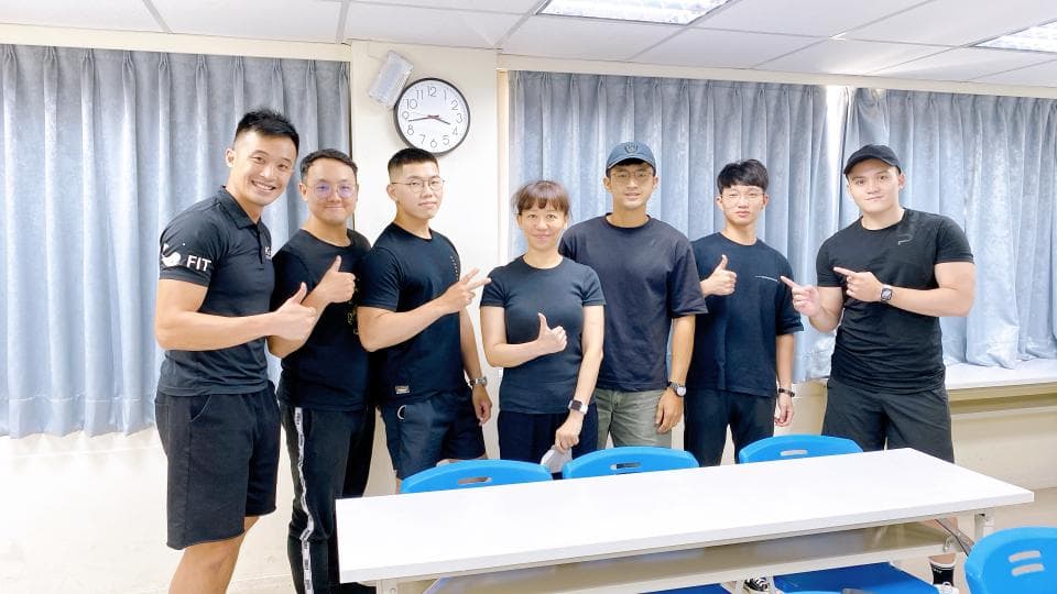 這張照片是七位男士在一個房間裡,他們站在白色的長桌旁,其中一位男士穿著黑色短袖上衣和黑色短褲,上面印有白色「FIT」字樣,他看起來是這群人的領袖。