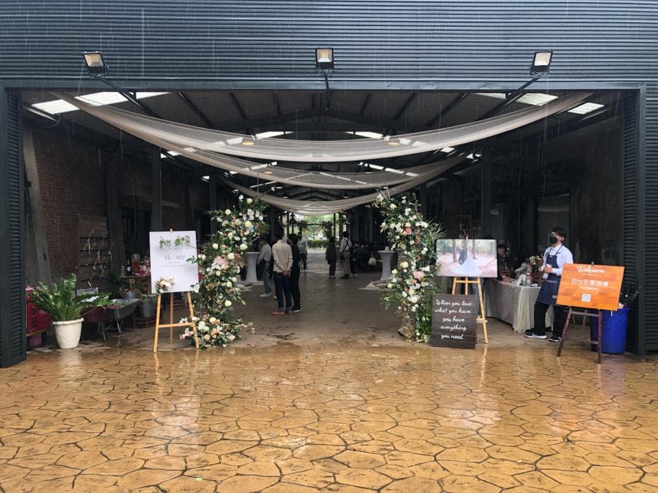 這張婚禮場地的照片是,在一個戶外空間裡,有兩個花拱門裝飾,前方有兩個木板寫著歡迎標語。