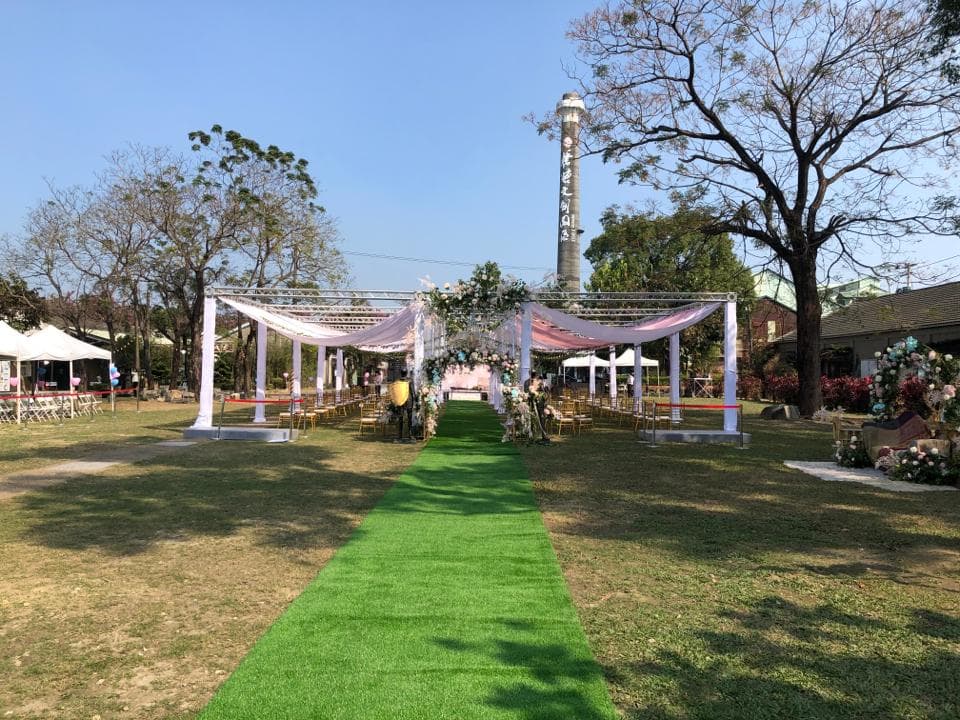 這張「戶外婚禮」的照片是,一個綠草地婚禮場地,有兩個白色帳篷和一個紅色的地毯,後面是一棵高聳的煙囪。