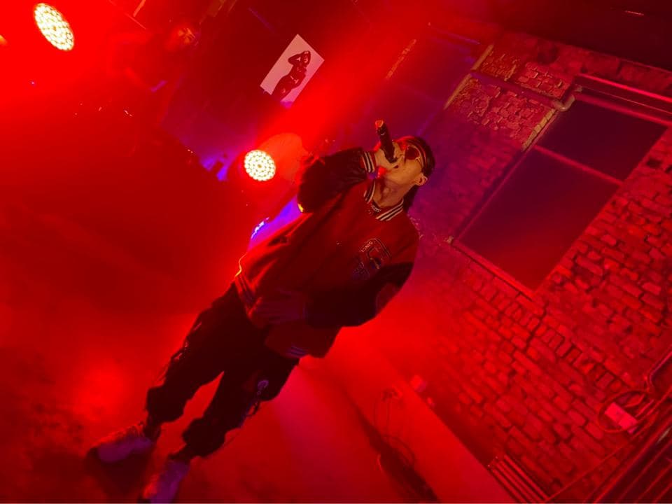 這張照片是一個歌手在紅色的燈光下唱歌。他戴著墨鏡,穿著一件紅色的外套和黑色的褲子。