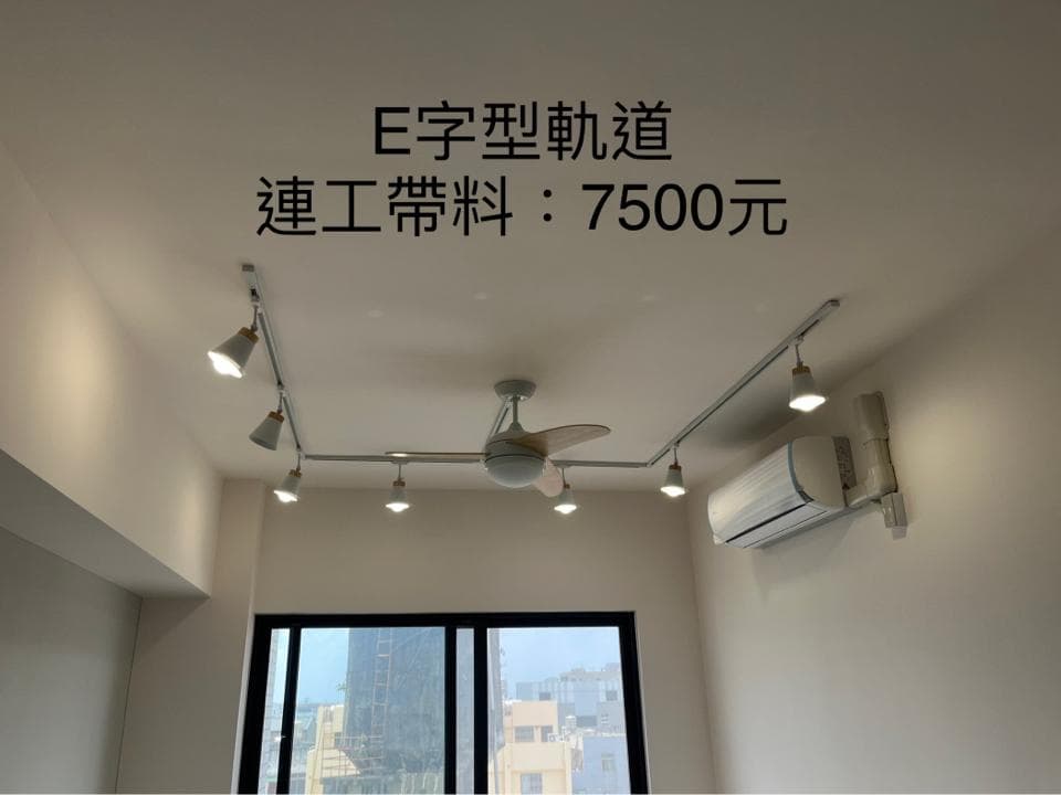 1) 照片顯示一個客廳,裡面有白色的天花板和牆壁,有一個白色的 E 型軌道燈,安裝在房間中間,一個白色的吊扇,安裝在天花板中央,一個白色的冷氣安裝在牆壁上,以及一扇落地窗,窗外是城市景觀