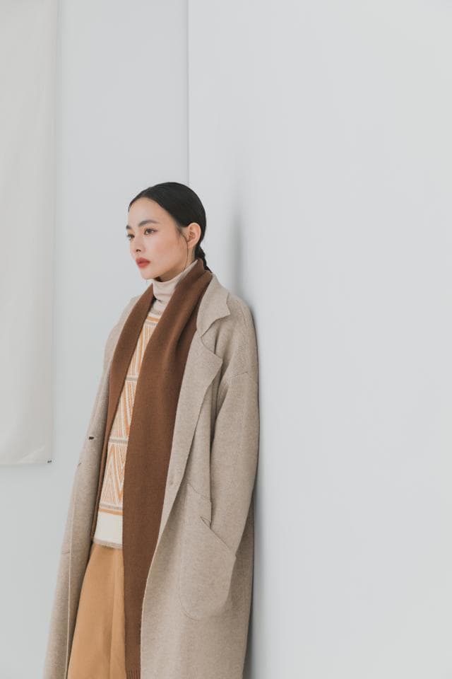 這張照片是一個穿著淺褐色大衣和深褐色圍巾的女孩,她側身站在牆邊,眼神望向遠方。