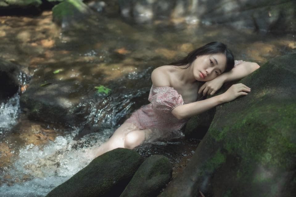 這張照片是一名女子躺在溪流邊的岩石上,她穿著粉紅色的蕾絲洋裝,看起來十分優雅美麗。