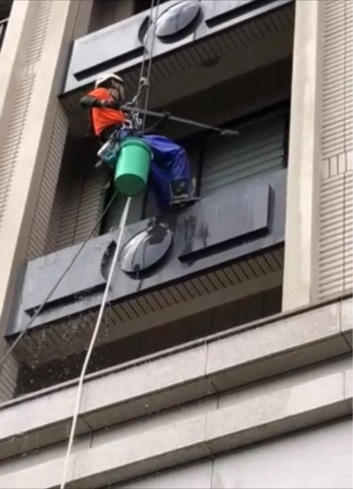 1) 照片顯示一名身穿橙色上衣和藍色褲子的男子,繫著繩索懸掛在建築物外,他手持水桶和清潔工具正在清洗窗戶