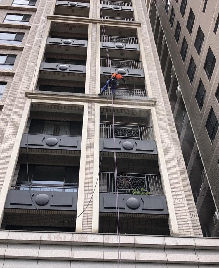 1) 照片顯示一名身穿橘色背心的男子,懸掛在繩索上,正在清洗大樓外牆的窗戶