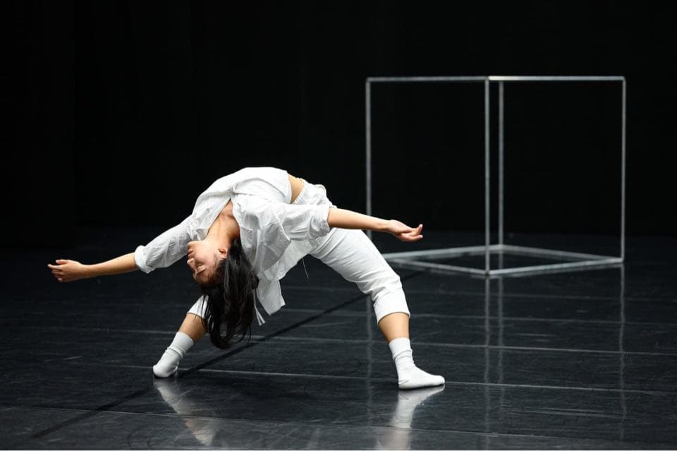 這張照片是一個穿著白色衣服的女性舞者,她正在表演一個優雅的舞蹈動作。她的身體像一條柔軟的線條,在空中優美地流動,呈現出極致的平衡和控制。她的一隻手伸向前方,另一隻手輕輕地觸碰著地面,彷彿要與地面的力量進行對話。她的動作充滿了力量和美感,讓人感受到舞蹈的魅力。