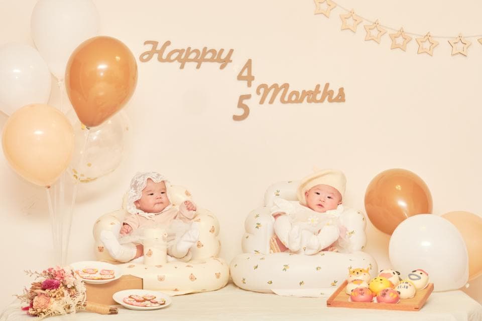 這張「雙胞胎寶寶慶祝四個月和五個月」的照片是兩個小嬰兒坐在可愛的嬰兒座椅上,背景佈置了氣球和星星串,看起來十分溫馨可愛。