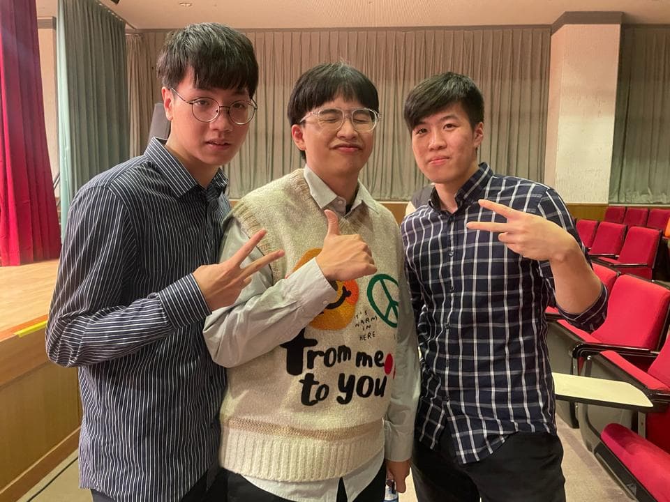 這張照片是三個人站在一起,他們都穿著休閒服裝,看起來像是朋友。他們背景是教室,牆壁上裝飾著窗簾。他們看起來很開心。