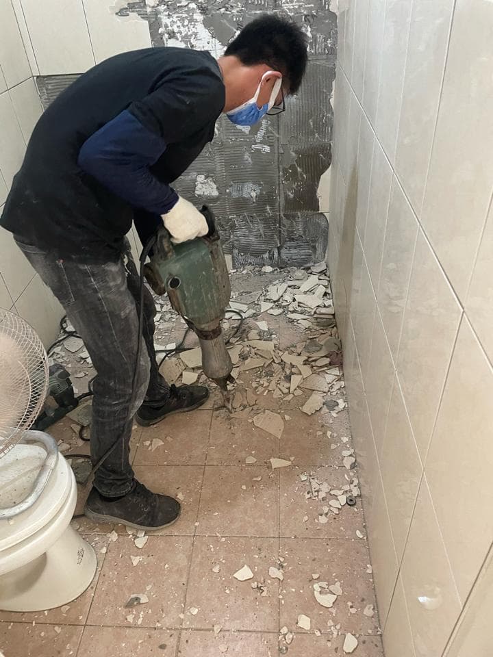 1) 照片中有一位男性在浴室裡用電鑽拆除牆壁,地上有許多瓷磚碎片,牆壁看起來是白色瓷磚牆