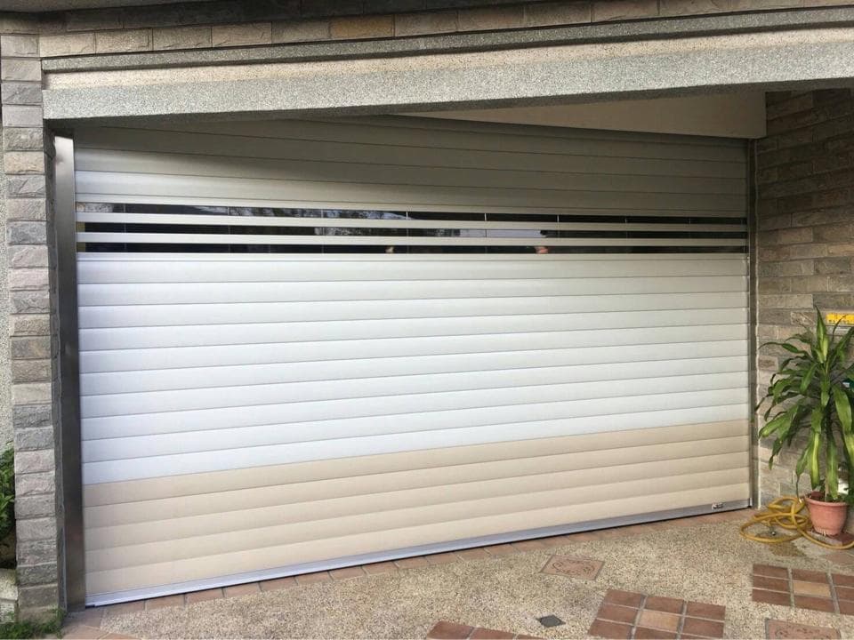 這張照片是一間房子外面,有一個電動捲門,電動捲門是白色的,上面有兩條灰色的線。