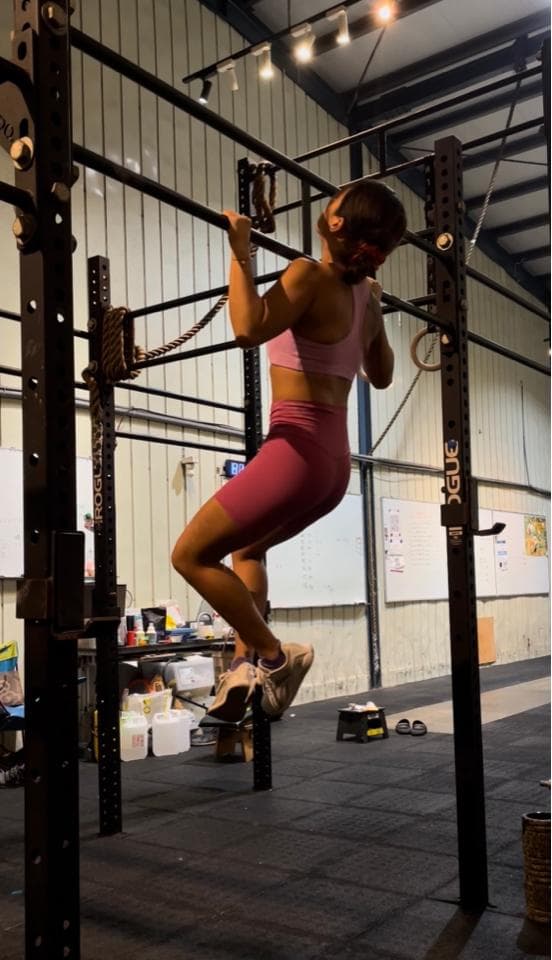 這張照片是一個女生在健身房做引體向上。她穿著粉紅色的運動服,雙手緊握著單槓,身體懸空,雙腳離地。她正在努力地向上拉,展現出堅定的意志和強健的體魄。