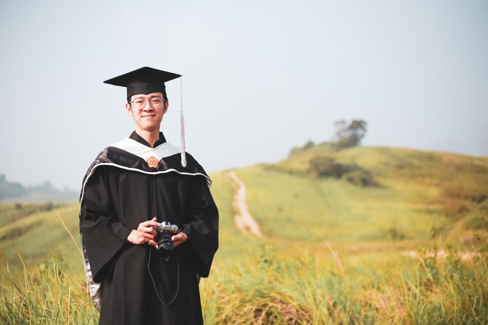 這張照片是一名身穿畢業服的年輕男性,他在一個綠色的草地上,手裡拿著一臺相機,他看起來很開心。