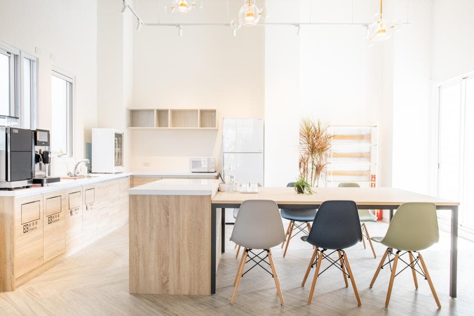 這張「辦公室」的照片是一間簡約明亮的空間,裡面有廚房、會議桌和幾張椅子。廚房有木質櫥櫃和白色家電,會議桌是木質的,上面放著幾盆綠色植物,椅子是黑色金屬腳架的。