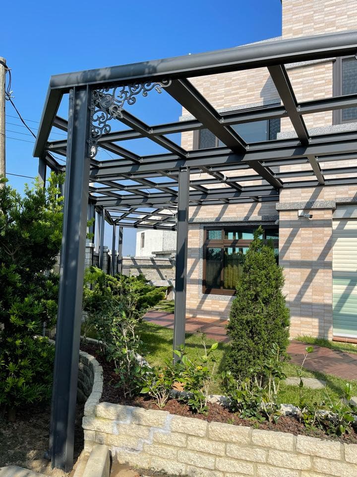 這張「戶外遮陽棚」的照片是一座鐵製的遮陽棚,棚架上裝飾著精緻的鐵花,並附有玻璃,讓整體看起來更美觀。遮陽棚旁邊還有一排石頭砌成的矮牆,牆上種植了綠色植物,增添了一份自然氣息。