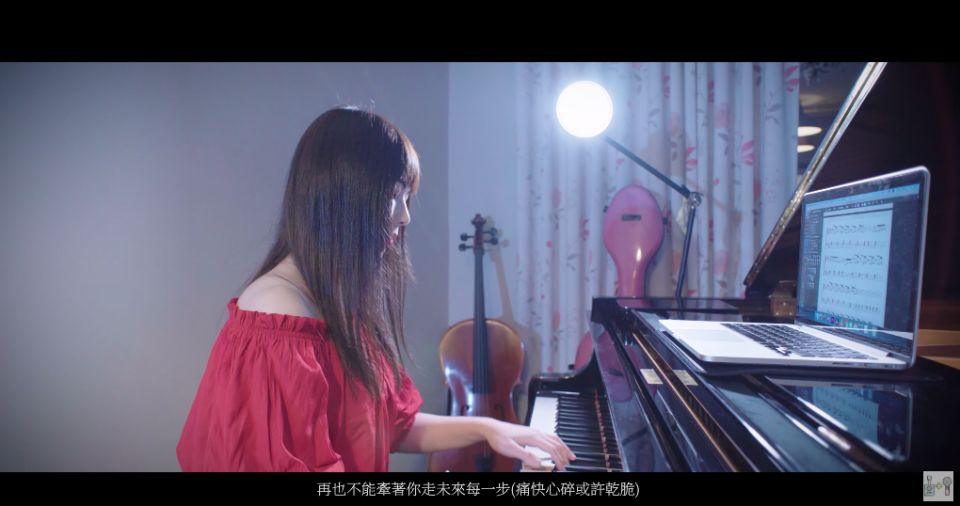 這張照片是一個年輕女子坐在鋼琴前彈奏的畫面。她穿著一件紅色的上衣,面前放著一臺筆記型電腦,顯示出音樂譜。房間裡還有一把大提琴和一盞燈。