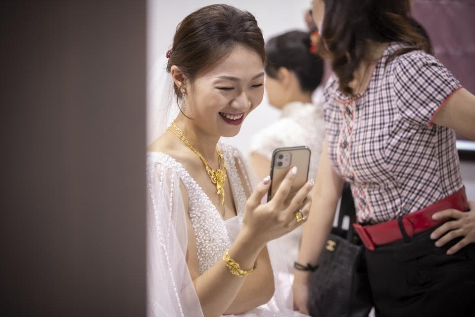 這張「新娘在婚禮前使用手機」的照片是,一位穿著婚紗的新娘正在看手機,她旁邊是她的朋友,她們看起來很開心。