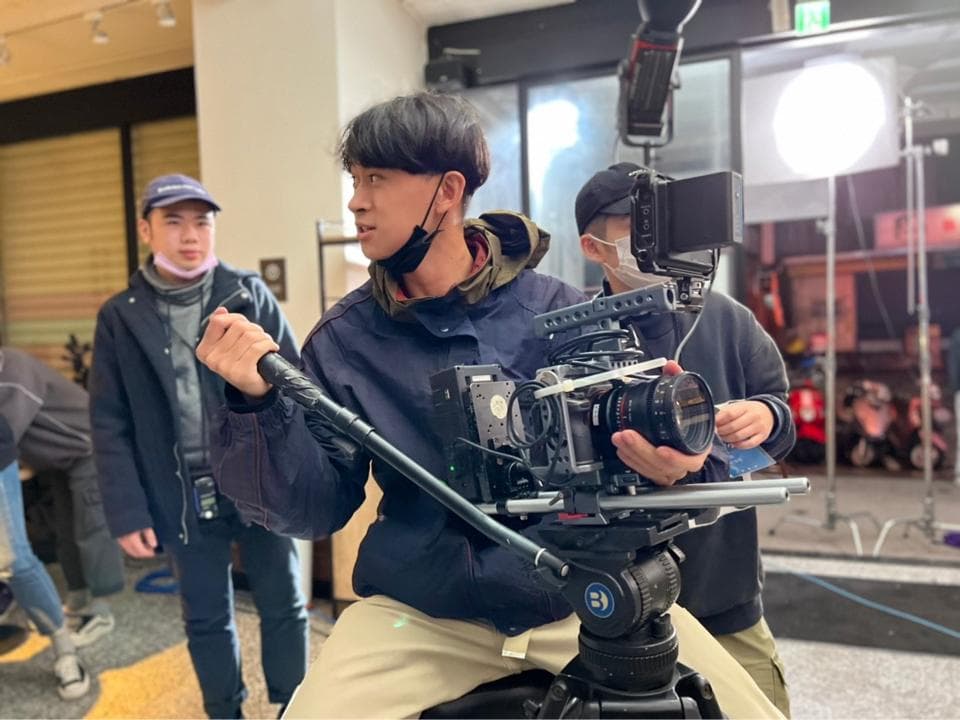 這張照片是一個攝影師正在使用專業攝影機拍攝。他專注地透過鏡頭觀察,手握著攝影機的握把,展現出專業的攝影態度。