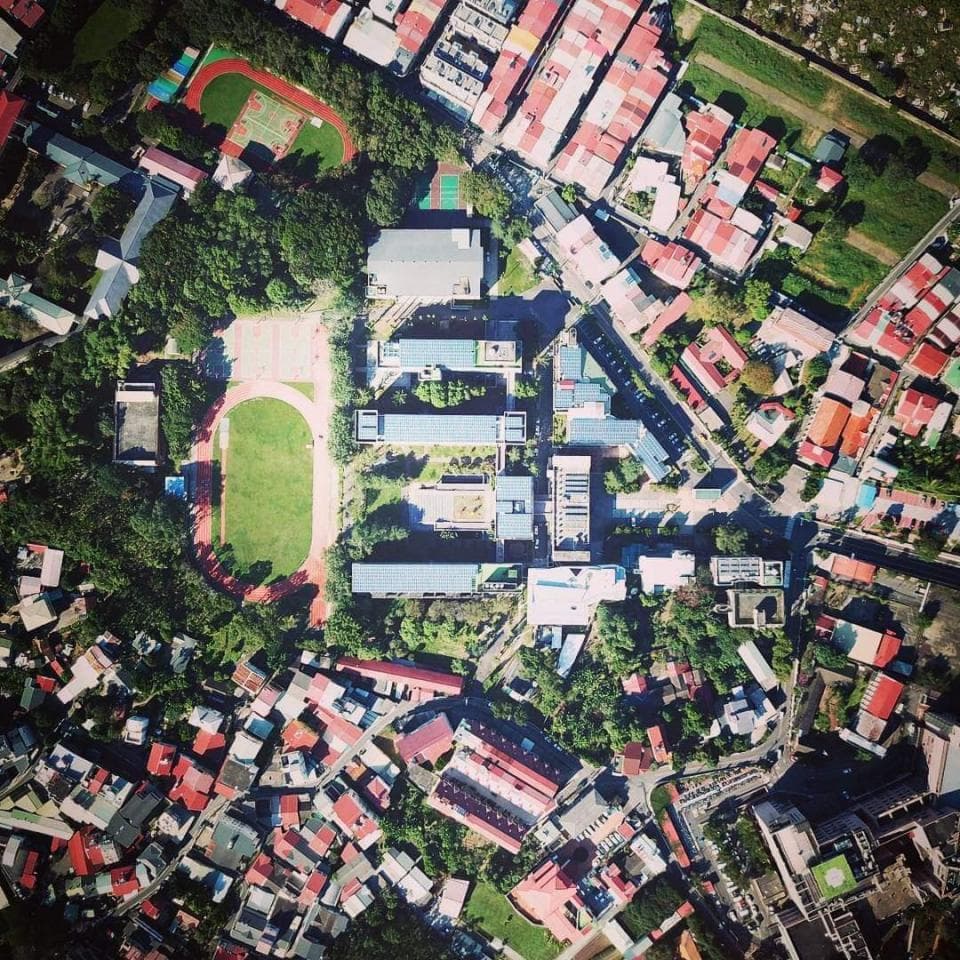 這張「學校操場」的照片是,學校操場旁有許多建築,操場旁邊有綠色的樹木。