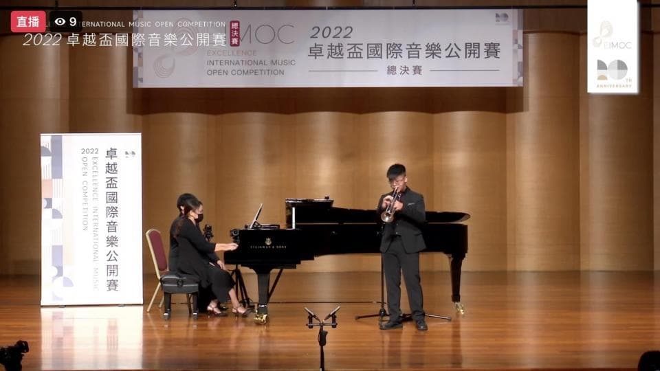 這張「音樂比賽」的照片是一名穿著黑色西裝的年輕人正在吹奏小號,而另一名穿著黑色連衣裙的女士正在彈奏鋼琴。他們正在舞臺上表演,背景是木質牆壁和一個標誌。