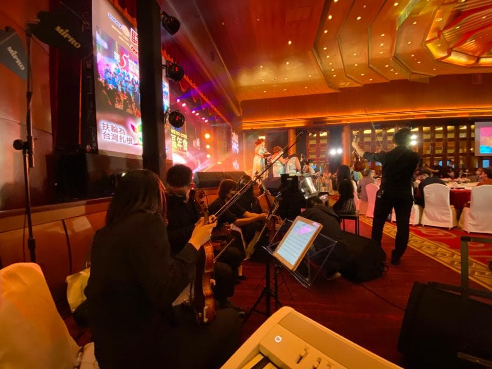 這張照片是婚禮現場,舞臺上有一位身穿白色禮服的女性正在演唱,臺下有幾位穿著黑色禮服的音樂家正在演奏。