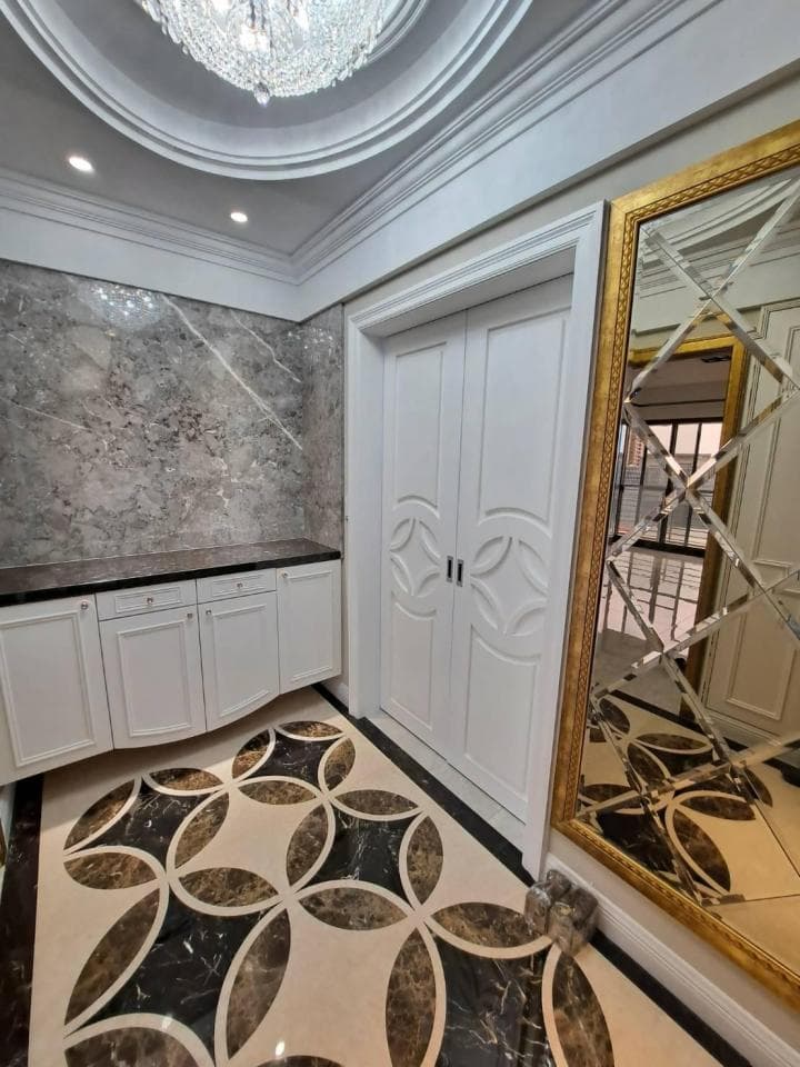 這張照片是客廳裡的一扇雙開門,門上雕刻著精美的花紋,門框旁的牆壁上裝飾著一面金色的鏡子,鏡子裡映照著大理石地板的花紋。