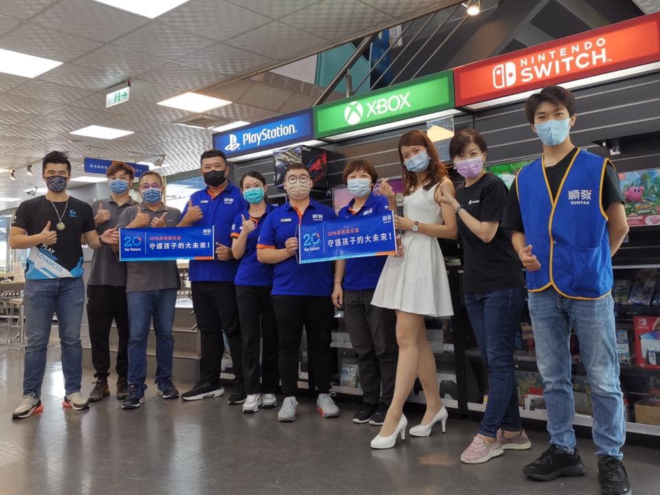 這張照片是幾位年輕人在一家電玩遊戲店舉辦活動,他們手持著「20週年慶祝活動」的看板,背景是各式各樣的電玩遊戲主機,以及琳瑯滿目的遊戲軟體。