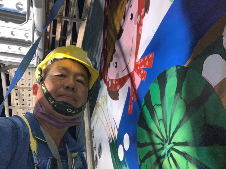 這張「一位戴著安全帽的工人站在一幅牆壁上的壁畫前」的照片是。壁畫上有農村場景,其中包含一個戴著紅帽子的女人、稻田和西瓜。工人穿著藍色的工作服,戴著口罩。