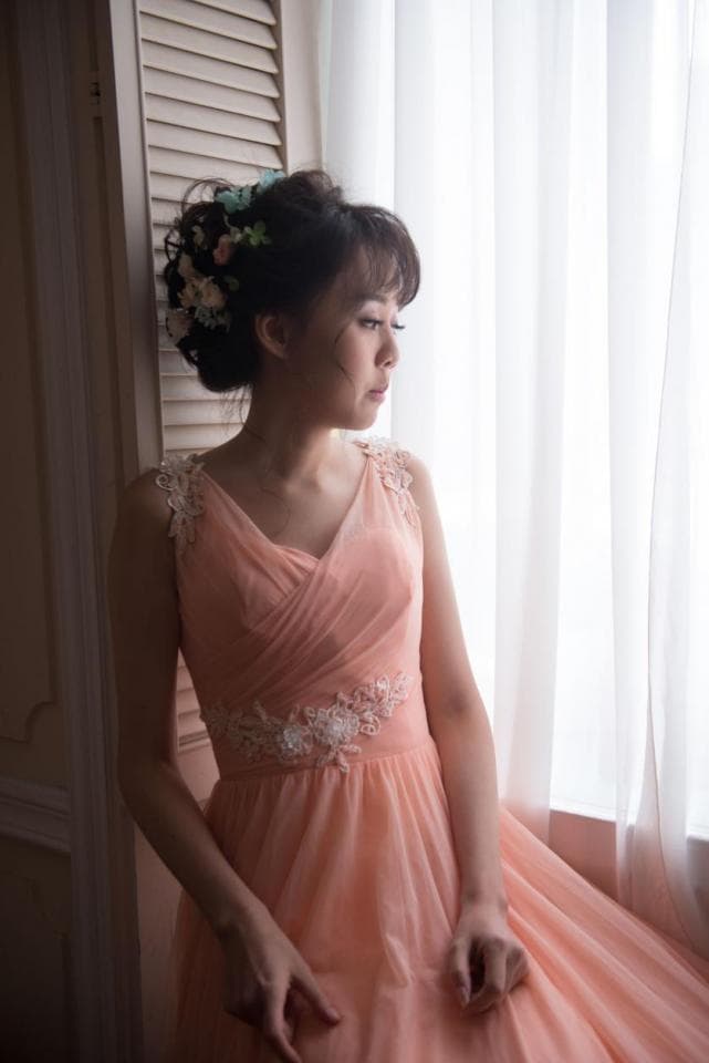 這張照片是一位穿著粉紅色連衣裙的年輕女子,她站在窗邊,凝視著窗外。她的頭髮上戴著花朵裝飾,看起來很優雅。