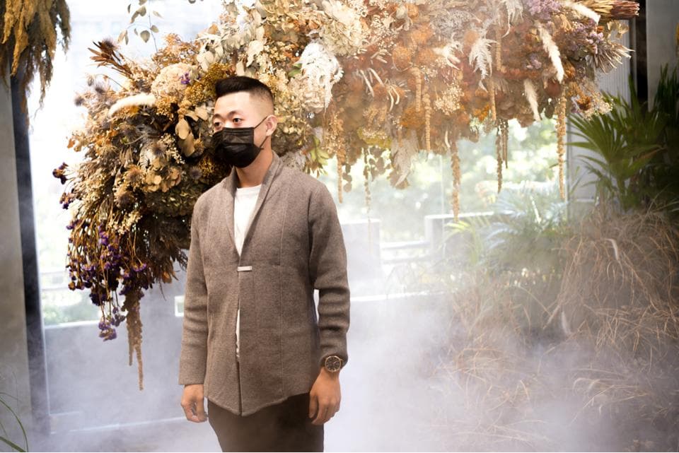 這張「一位戴著口罩的男人站在乾燥花叢下」的照片是,他穿著棕色毛衣,背景有綠色植物。