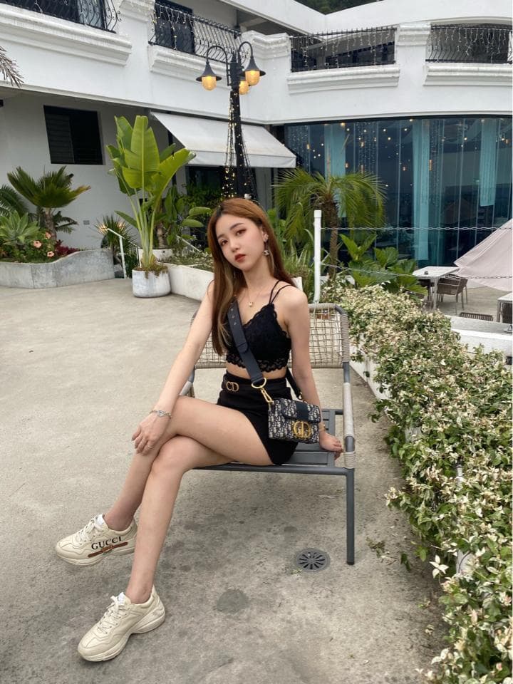 這張照片是一個年輕女性坐在戶外長椅上的照片。她穿着黑色的短上衣和短裙,手上拿着一個時尚的包包。她看起來很時尚,也很放鬆。