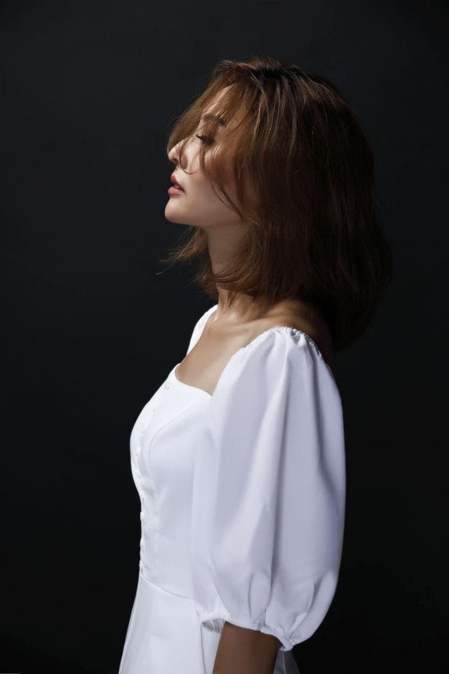 這張「一位穿著白色連衣裙的女性」的照片是。她有一頭棕色頭髮,閉著眼睛,看著鏡頭。她穿著一件有泡泡袖的白色連衣裙,站在黑色的背景前。