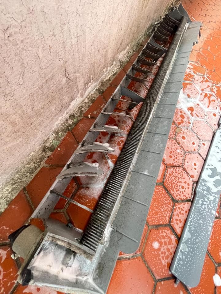 這張照片是一個灰色的金屬排水溝,它被安裝在牆壁和紅磚地面之間。排水溝上有許多小孔,可以幫助水流走。排水溝的表面看起來很舊,上面佈滿了污垢。