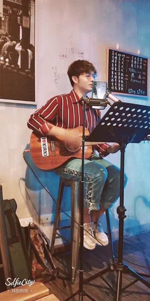 這張照片是一個年輕男子坐在高腳椅上彈吉他的畫面,他穿著紅白條紋襯衫,戴著眼鏡,看起來很認真地在表演。