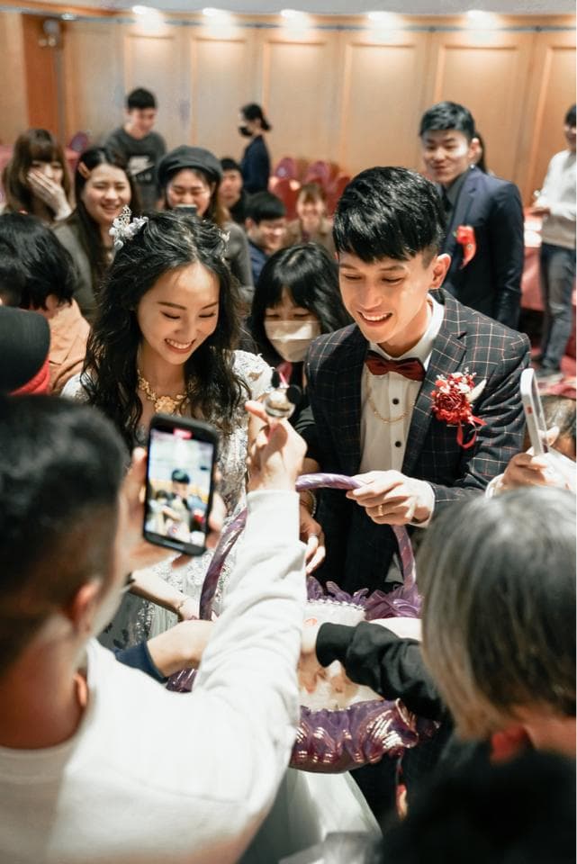 這張照片是一對新人在婚禮上進行傳統儀式,新郎正從一個籃子中拿出物品,而新娘則在一旁看著,他們身穿喜慶的服裝,臉上洋溢著幸福的笑容。