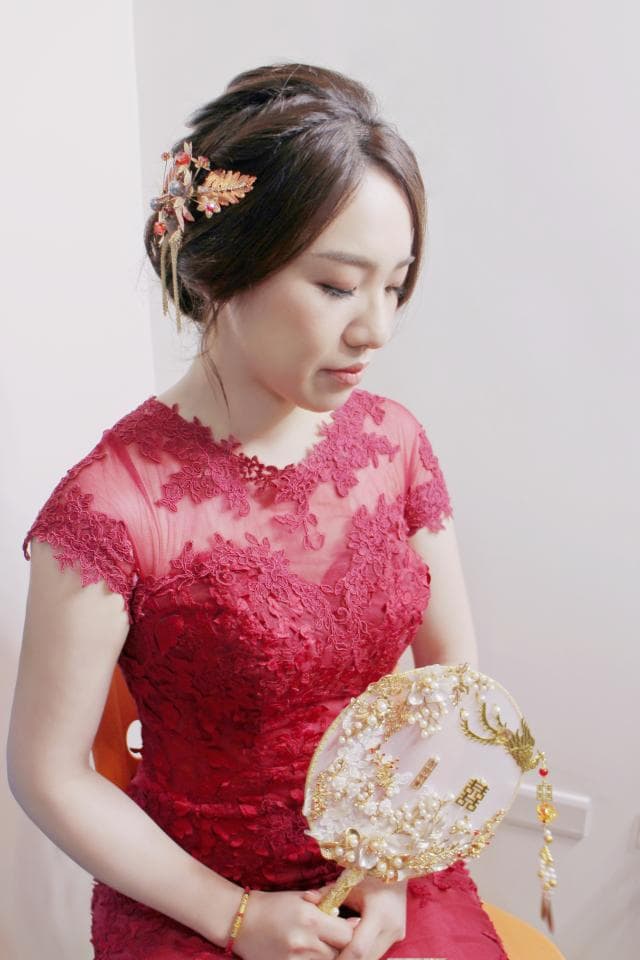 這張照片是一位身穿紅色蕾絲禮服的新娘,她手持著精緻的金色鑲珠扇子,看起來既優雅又喜氣。