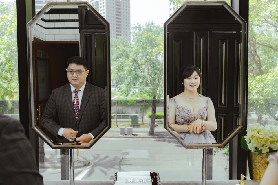 這張「鏡子」的照片是兩個人站在鏡子前,一個穿著西裝,一個穿著洋裝,背景是窗戶和綠色植物。